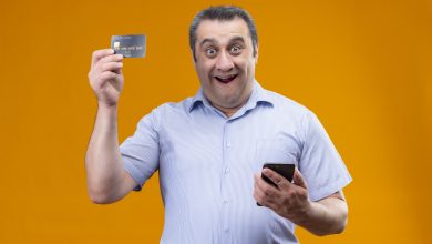 cheerful and positive middle age man wearing blue stripped shirt holding mobile phone and showing credit card while standing