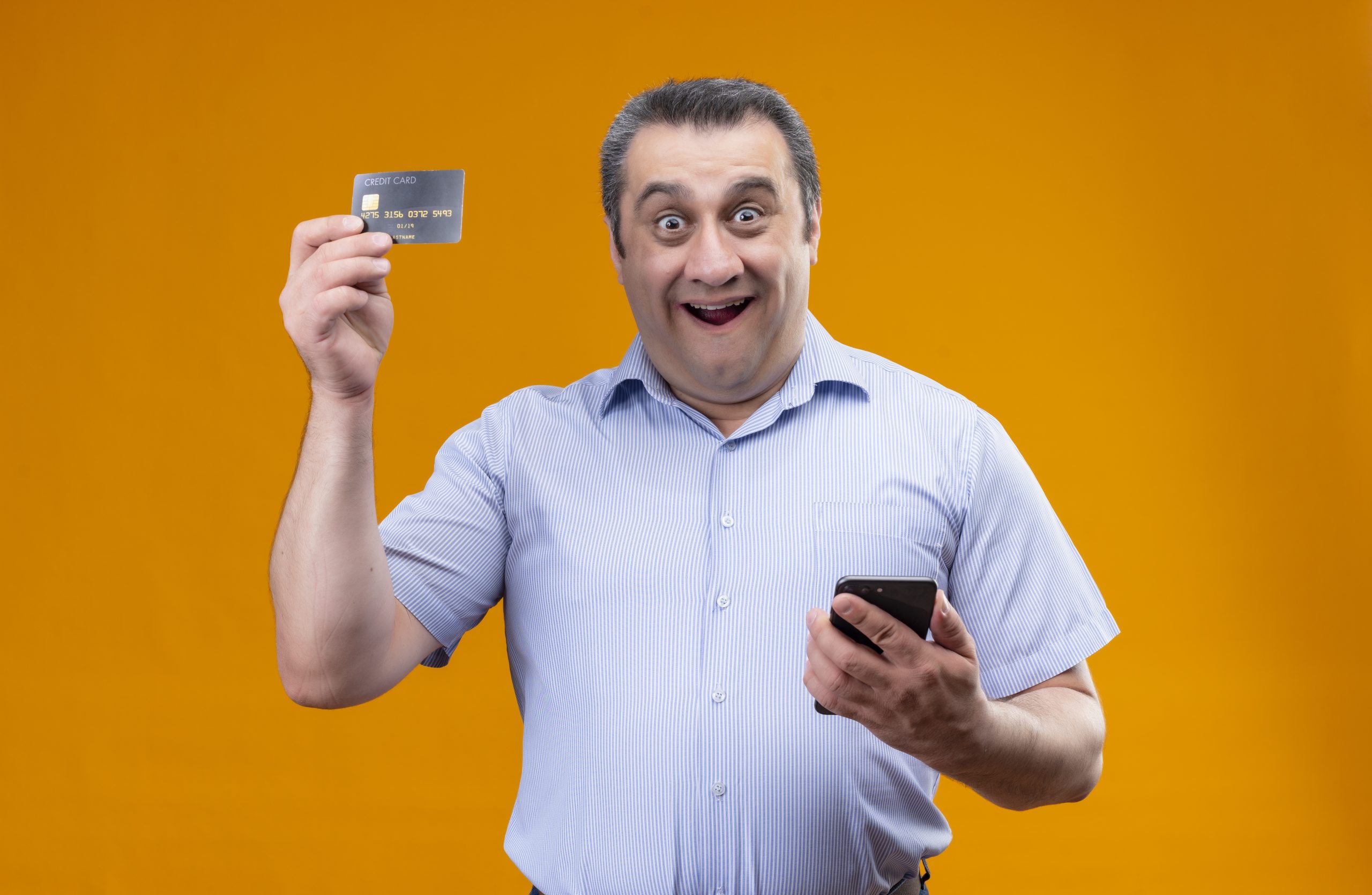 cheerful and positive middle age man wearing blue stripped shirt holding mobile phone and showing credit card while standing scaled