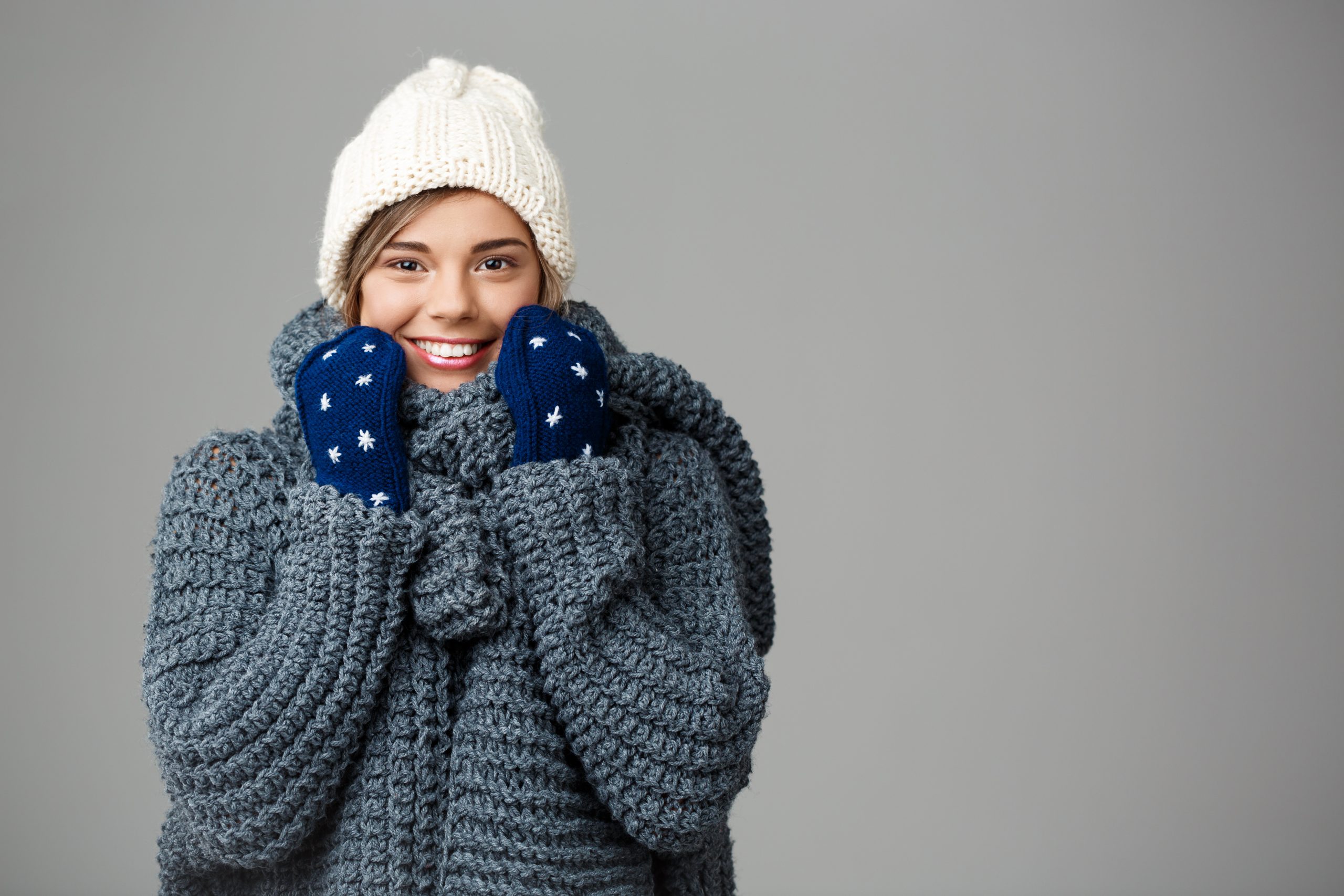 young beautiful fair haired woman in knited hat sweater and mittens smiling on grey scaled