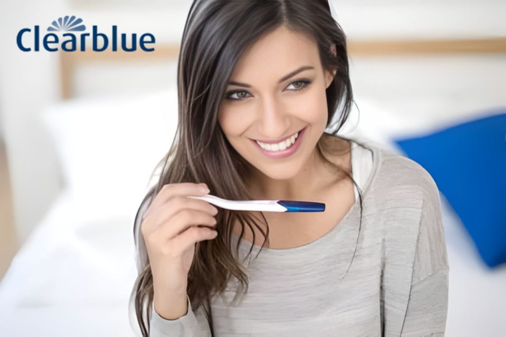 Mulher sorridente segurando um teste de gravidez Clearblue, representando a verificação dos sintomas da gravidez de forma prática e confiável.