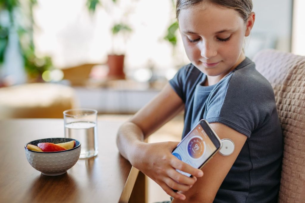 Criança usando sensor de glicose no braço e acompanhando as leituras no celular, demonstrando como aplicativos de monitoramento de glicose facilitam o controle do diabetes.