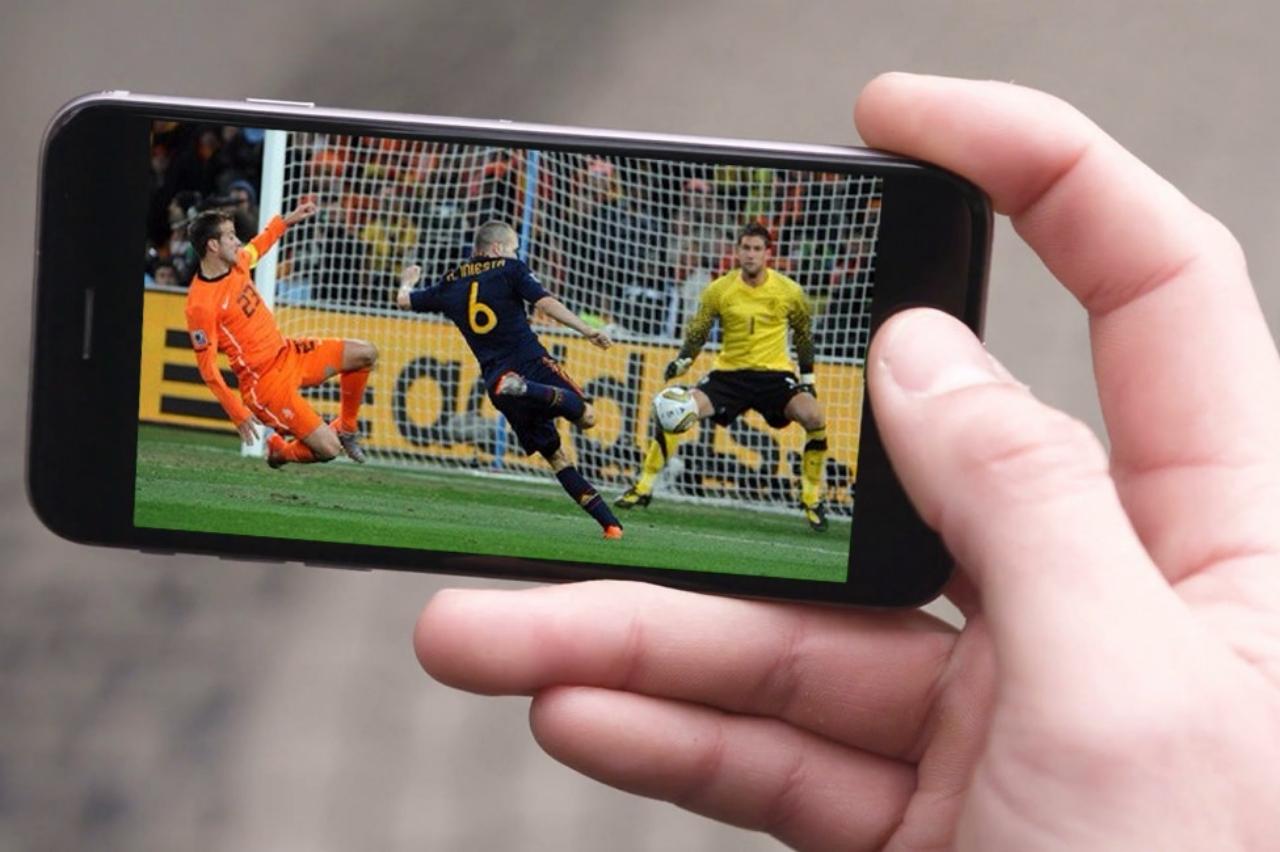 Pessoa segurando smartphone com partida de futebol ao vivo na tela, representando o uso de aplicativos para assistir futebol ao vivo pelo celular com praticidade e qualidade.